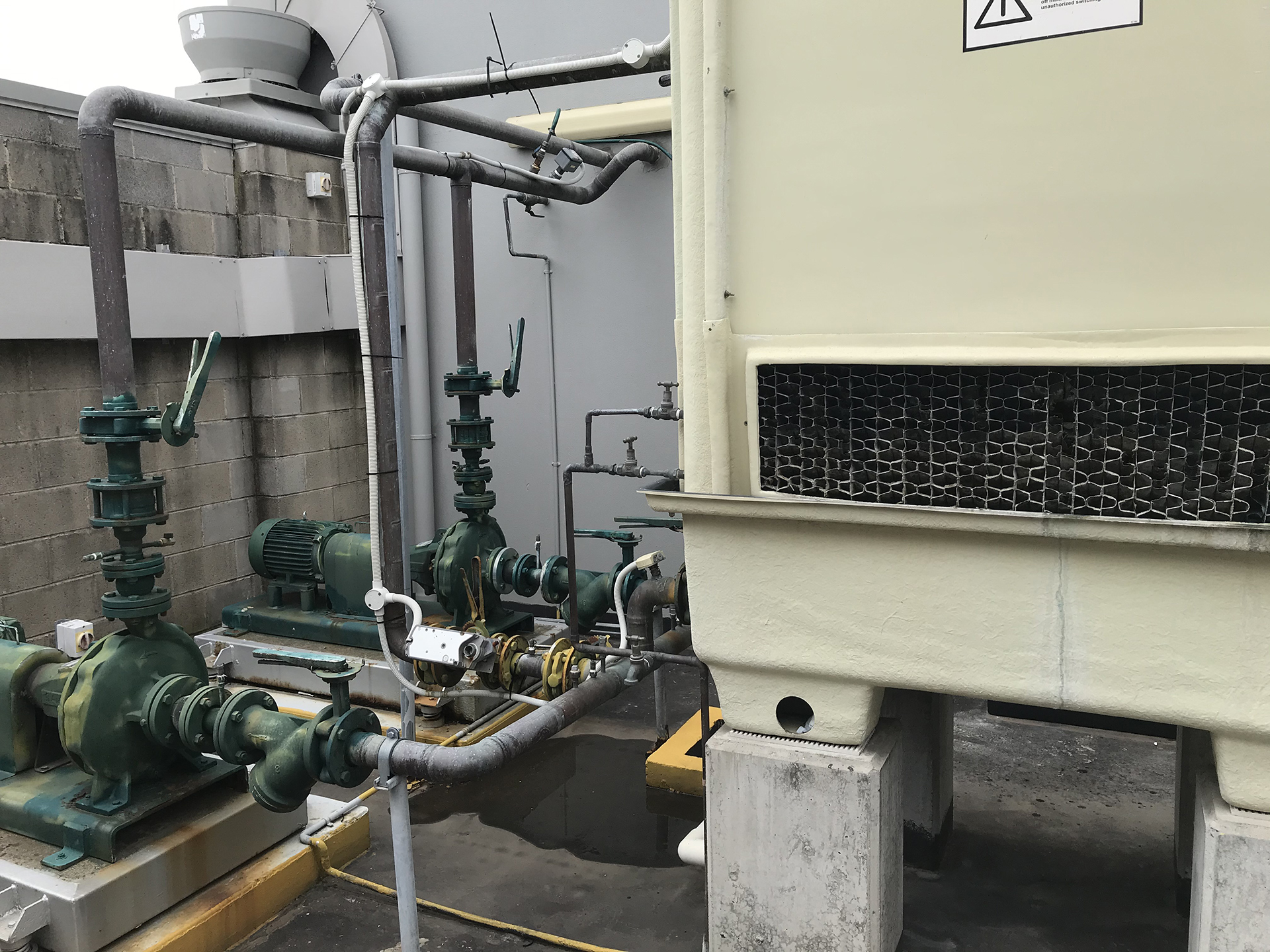 A commercial rooftop cooling tower installation in Leichhardt, Sydney, serving a large HVAC condenser water system. The image shows a cooling tower mounted on a steel platform with two condenser water pumps located adjacent to the tower and connected via insulated condenser water pipework. The pumps are configured in a lead/lag arrangement, allowing automatic duty and standby operation to improve reliability, reduce wear, and maintain continuous building cooling. Large supply and return condenser water pipes, isolation valves, strainers, and balancing valves are visible throughout the installation. The rooftop plant area is well maintained, with mechanical services infrastructure, safety handrails, electrical controls, and building services equipment visible in the background. The system represents a typical commercial building cooling tower and condenser water pumping arrangement designed for efficient heat rejection and energy performance in Sydney's commercial property sector.