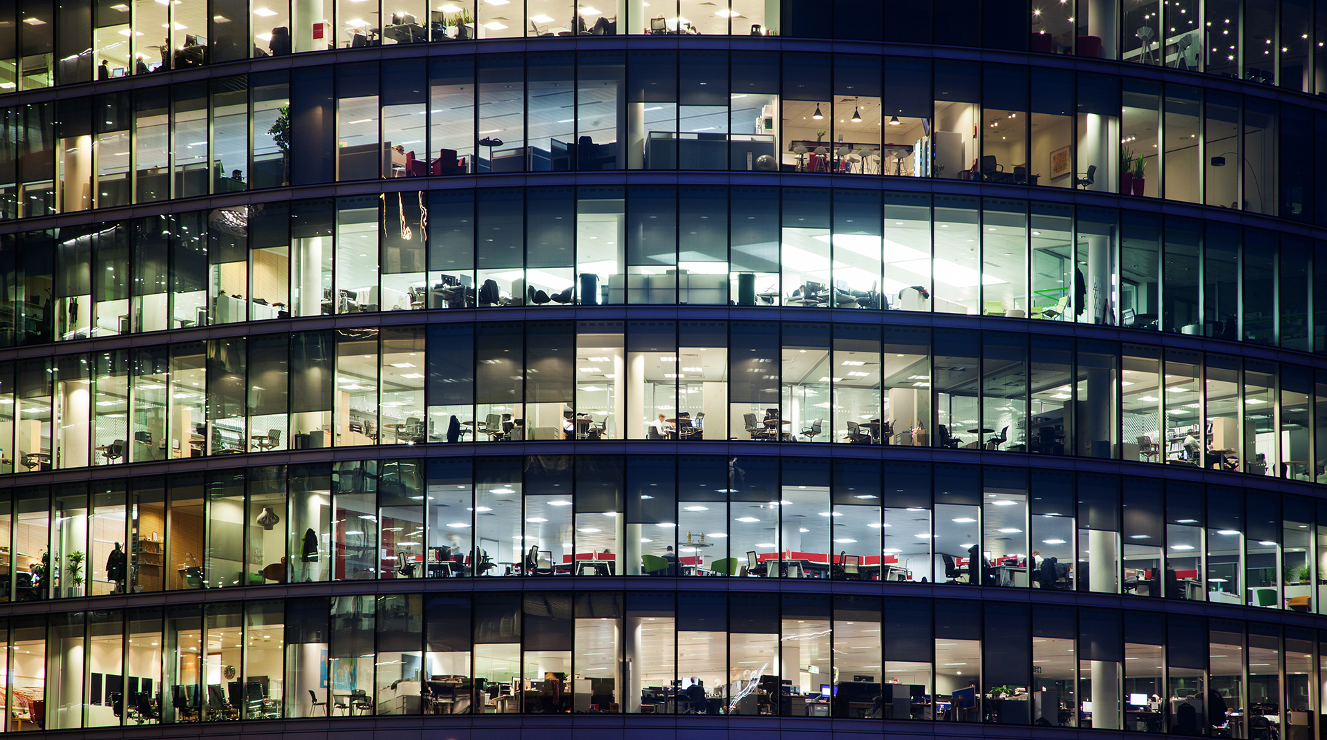 Sydney building fcade with lights on in all the windows where office furnitures and equipment can be seen with the humans, about 8 levels of windows