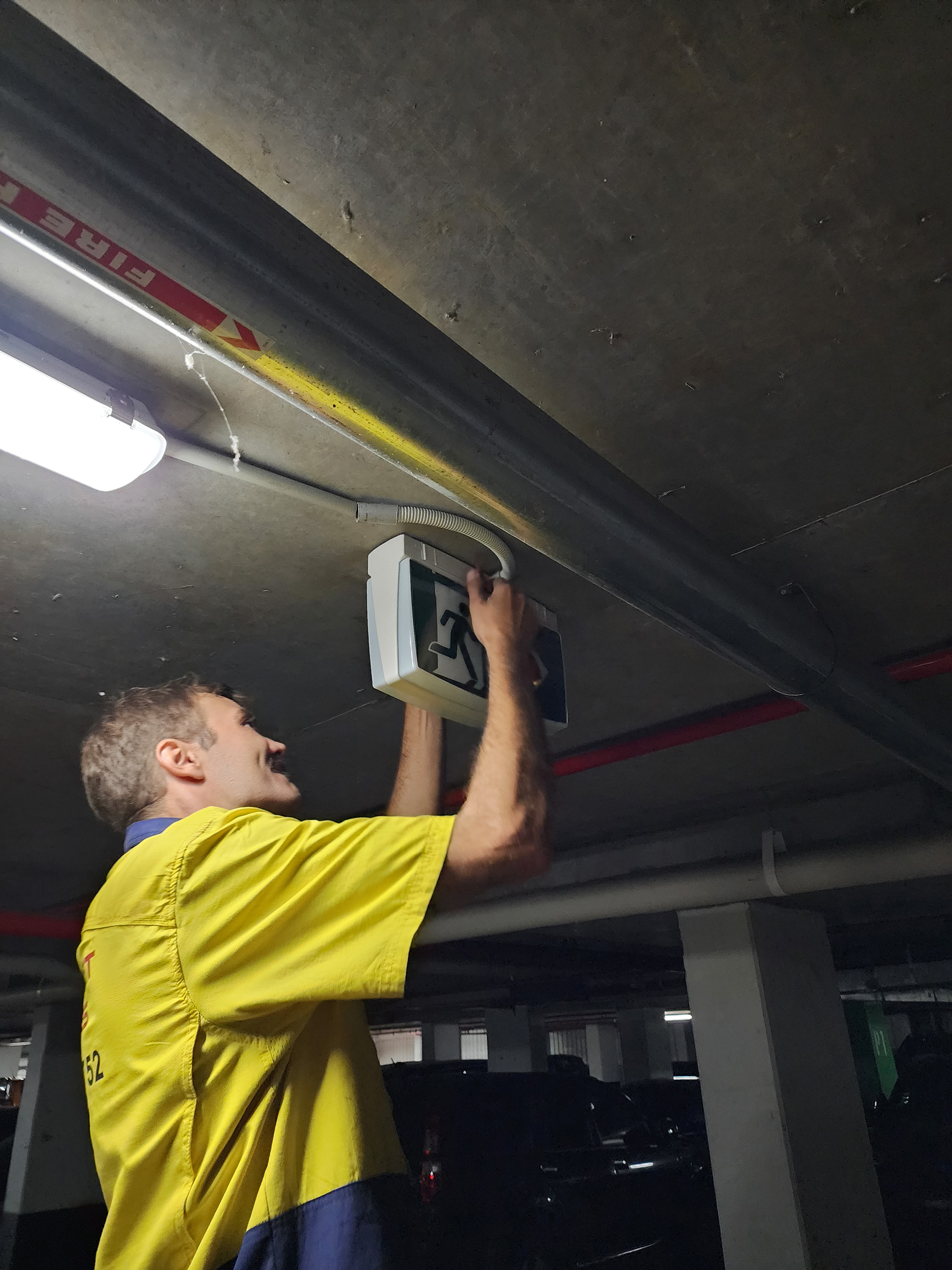 WR8Tech technician servicing an emergency exit light in a commercial basement car park as part of a lighting control and maintenance program.