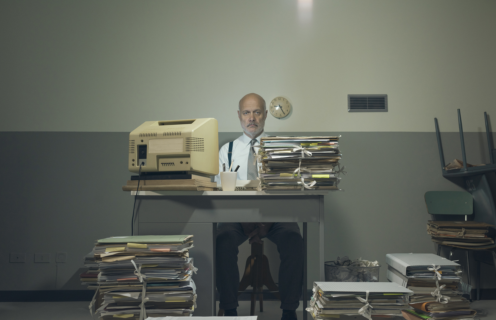 A manager sitting behinid a very old CRt computer screen with heaps of paper work, implying what it was like in the 1080's and 90's